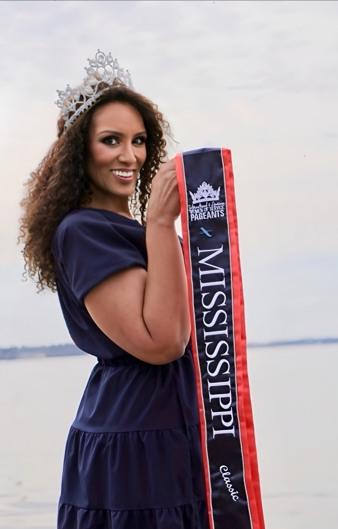 Woman wearing a crown and holding a “Mississippi Classic” pageant sash while smiling outdoors near the water.